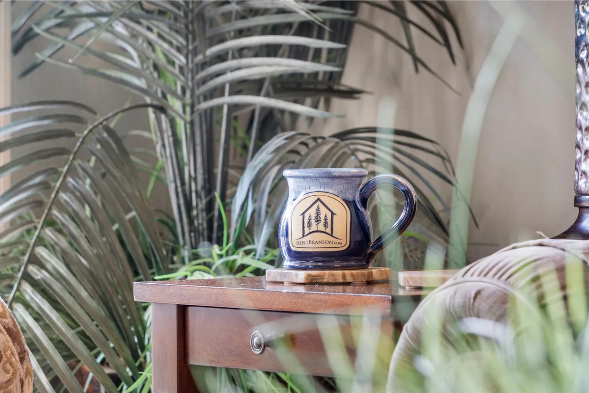 A rustic wooden table topped with a ceramic mug, surrounded by green plants and a softly glowing lamp. The scene feels cozy and peaceful, highlighting quiet mornings or evenings spent relaxing with a warm beverage.