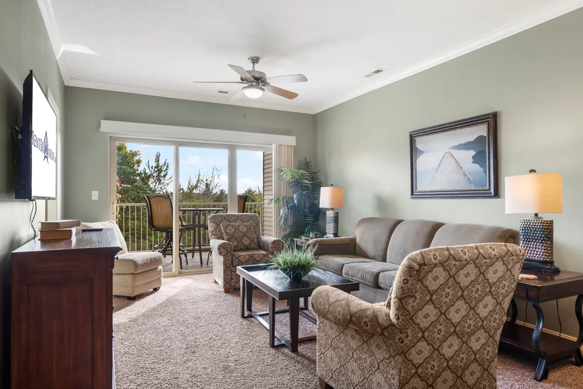 A comfortable living room setup with a sofa, coffee table, and armchairs facing a window that frames an inviting view. Subtle natural lighting suggests the presence of a balcony or outdoor area just beyond the glass.