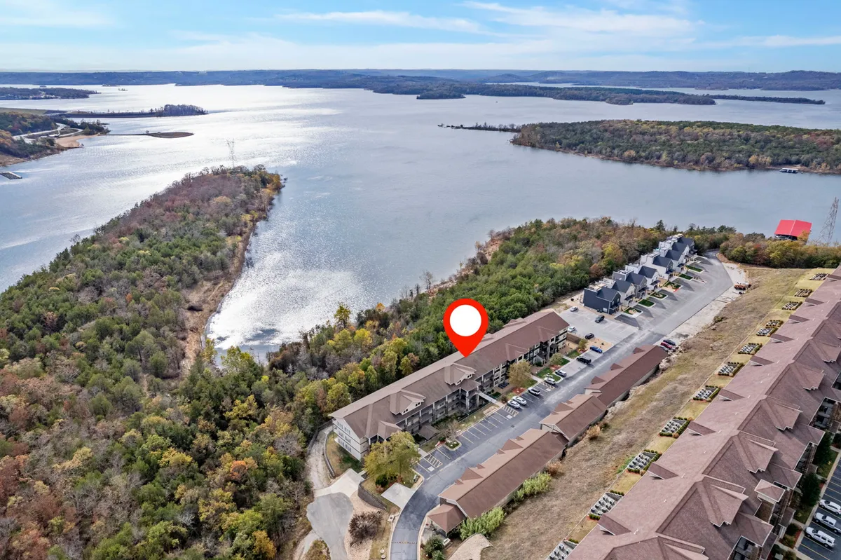 The image showcases the exterior of a waterfront condo surrounded by lush greenery, with neighboring buildings and a parking area visible. The scene is serene, capturing sunset views over the water and hinting at nearby opportunities for boating, hiking, and wildlife spotting.